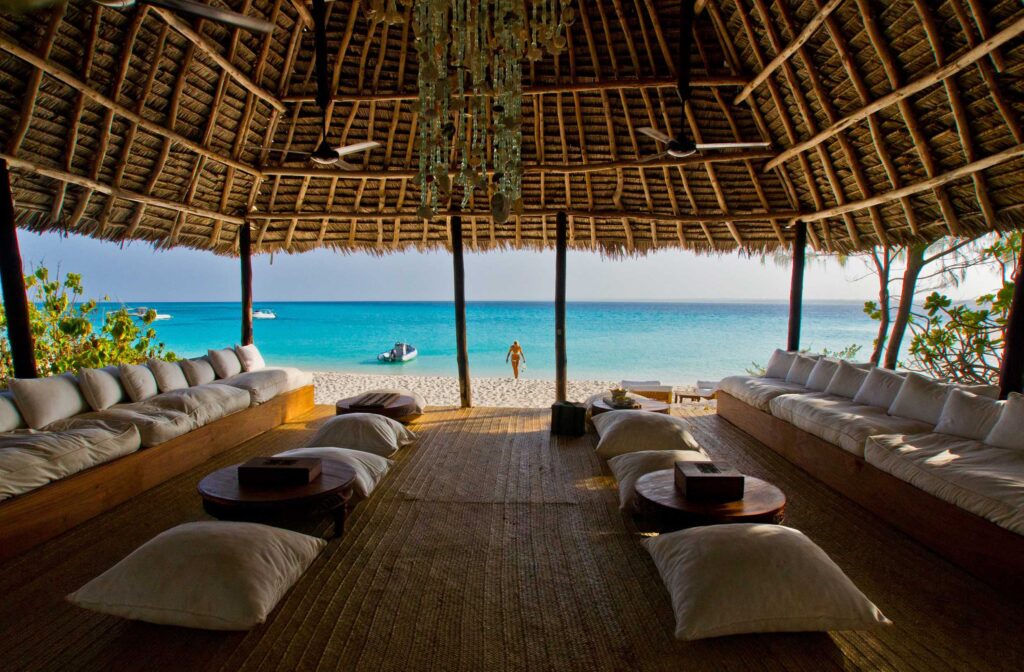 Matrix Safaris A thatched-roof open-air lounge with cushioned seating overlooks a sandy beach and turquoise ocean. A person stands at the shore, and a boat floats on the water in the distance.